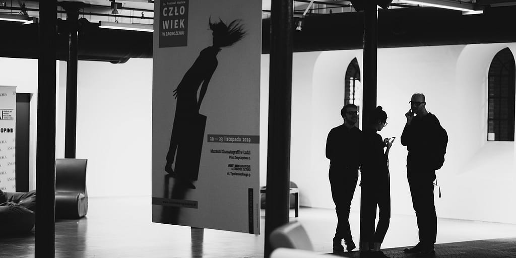 ALT - Sala o białych ścianach. Po lewej zawieszony plakat festiwalu, po prawej sylwetki trojga osób