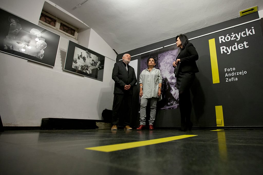 ALT - Sala wystawowa. Na ścianach fotografie. Na ich tle stoją trzy dorosłe osoby zwrócone twarzą do siebie.