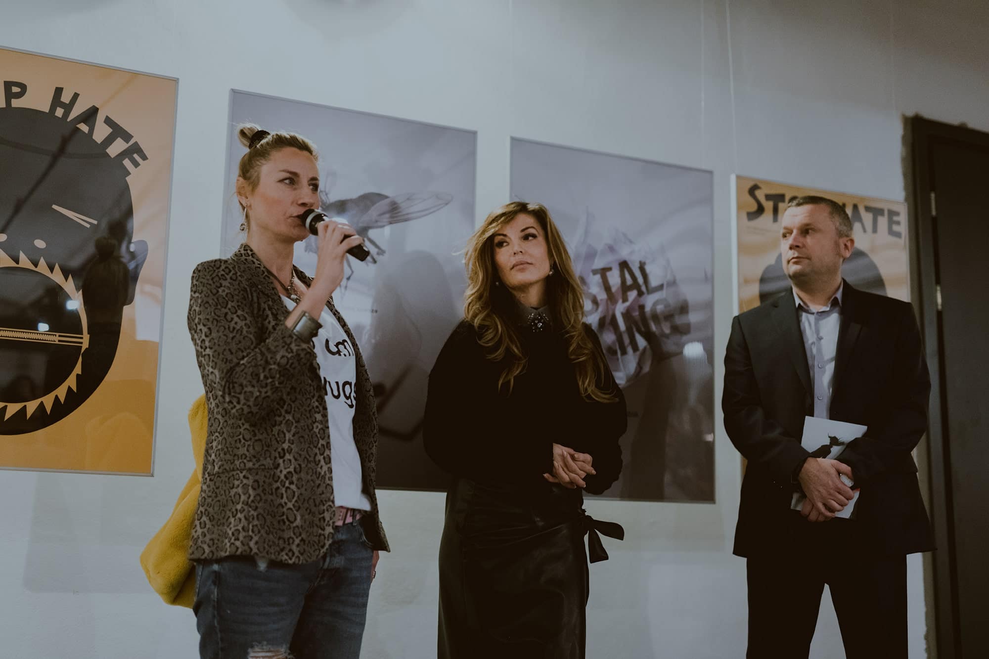 ALT - Dwie kobiety i mężczyzna stoją na tle galerii plakatów. Jedna z nich trzyma mikrofon przy ustach.