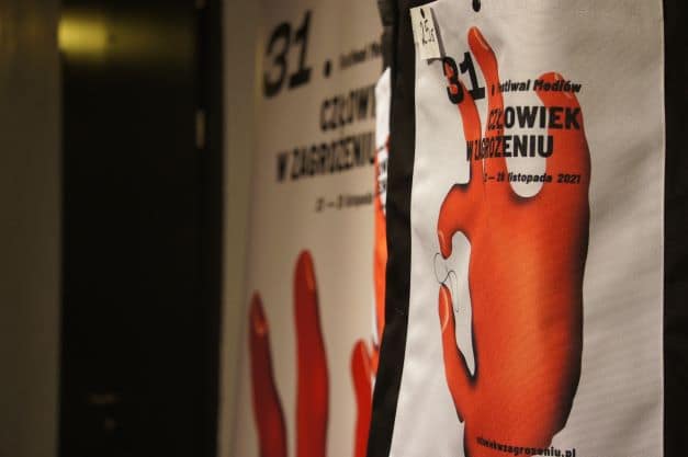 ALT - Zdjęcie wiszącego plakatu. Rozmazana i zniekształcona czerwona dłoń. Napis: 31. Festiwal Mediów Człowiek w Zagrożeniu.