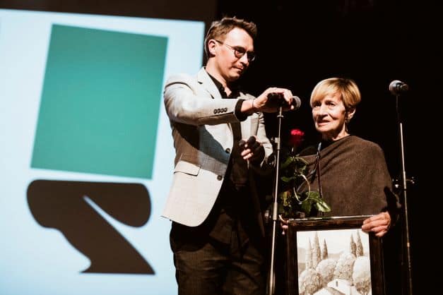 ALT - Mężczyzna przytrzymuje mikrofon, do którego mówi kobieta. Kobieta trzyma w rękach obraz i różę.