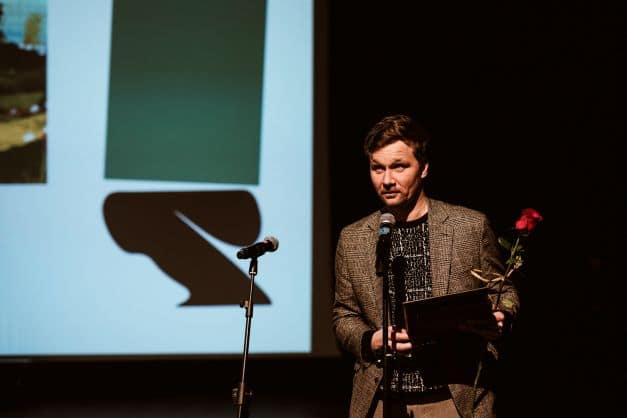 ALT - Mężczyzna w marynarce stoi przed mikrofonem. Pokazany z przodu, od pasa w górę. W tle ekran z logotypem Festiwalu.
