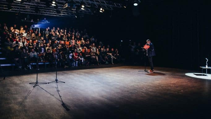 ALT - Sala widowiskowa. Na pierwszym planie drewniana podłoga. Na drugim stoi mężczyzna przy mikrofonie. W tle siedzi publiczność.